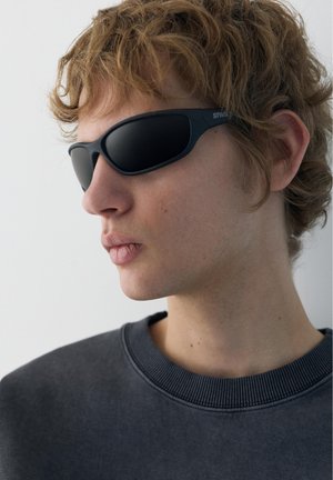 Mlada osoba s kovrčavom svijetlosmeđom kosom, nosi crne sunčane naočale s omotavanjem i tamnosivu majicu s okruglim izrezom, okrenuta prema lijevo na bijeloj pozadini.