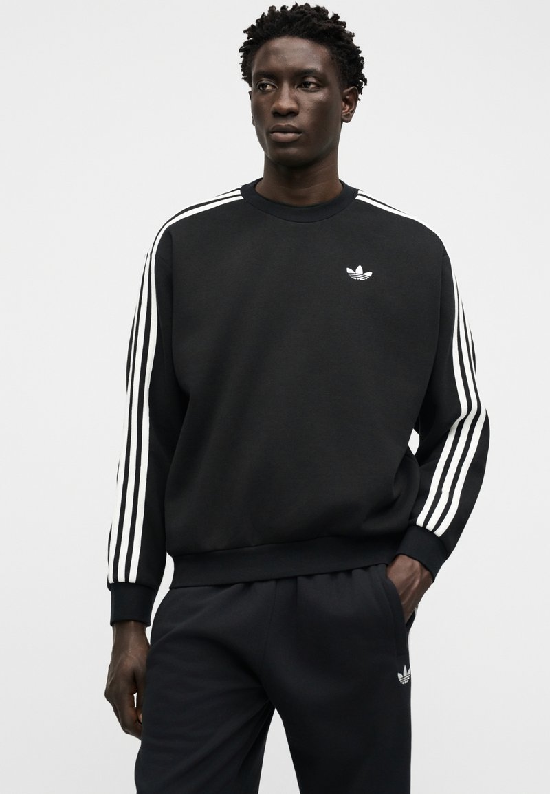 Man wearing black Adidas sweatshirt and pants with white three-stripe detailing and logos, posing with one hand in pocket.