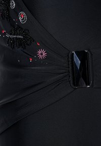 Sort stof med plisseringer holdt sammen af en blank sort rektangulær spænde, dekoreret med små broderede blomster og perlearbejde i hvid, rød og lyserød.