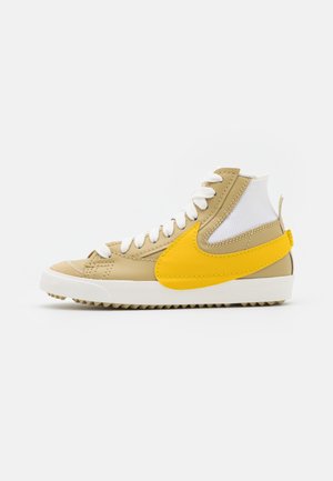 Beige hoge sneaker met witte veters, witte geribbelde enkelboord, gele Nike swoosh en witte zool met gestructureerd profiel.