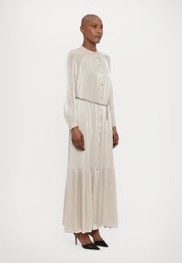 Robe longue en argent avec un design à boutons, en tissu plissé et avec un ourlet en étages. Elle présente une ceinture fine et des manches longues, assortie à des talons noirs.