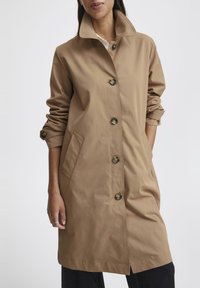 Cappotto trench khaki con colletto alto, chiusura a bottoni, due tasche laterali e maniche arrotolate con accenti a bottone. Texture liscia e opaca.