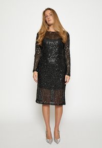 Schwarzes Paillettenkleid mit langen Ärmeln und transparenten Einsätzen, mit Rundhalsausschnitt und knielangem Schnitt, kombiniert mit silbernen High Heels.