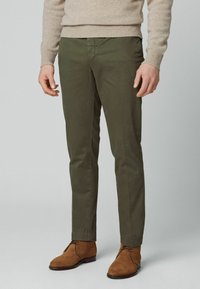 Homme portant un pantalon slim olive, un pull beige et des bottines à lacets en daim marron, debout sur un fond gris uni.