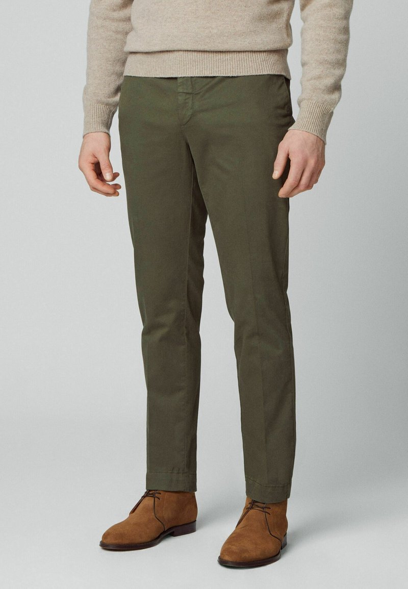 Homme portant un pantalon slim olive, un pull beige et des bottines à lacets en daim marron, debout sur un fond gris uni.