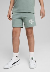 Pantalones cortos de algodón verde claro con cintura elástica, que presentan un logo blanco de "PUMA sports club". Combinados con zapatillas blancas y calcetines hasta el tobillo.