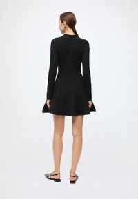 Black long-sleeve dress with a flared skirt, made of textured fabric, featuring a collar and back-zip closure, styled with flat black shoes.