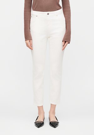 Dolná časť tela osoby oblečenej v bielych priliehavých nohaviciach po členky, hnedom dlhorukávniku a čiernych špičatých balerínach s remienkami.