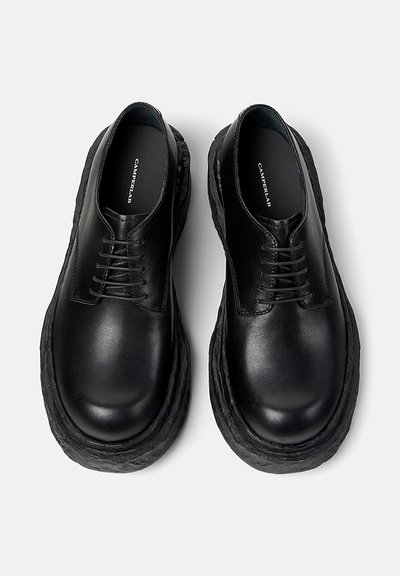 Pari mustia nahkaisia nauhallisia kenkiä, joissa paksut kuvioidut pohjat, kuvattuna ylhäältä vaaleanharmaalla taustalla.