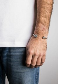Sidabrinis ir baltas apyrankė dėvima ant rankos; sidabras turi tekstūruotą raštą su sagtimi, o baltas dirželis yra lygus ir lankstus.