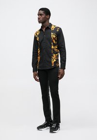 Camisa negra de botones con un patrón abstracto amarillo en un lado; combinada con pantalones negros y zapatillas negras con detalles blancos.