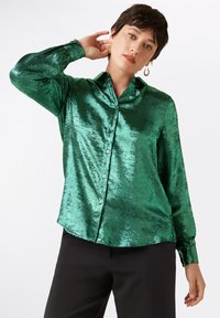 Groene satijnen blouse met een knoopsluiting aan de voorkant, lange mouwen en een klassieke kraag. De stof heeft een glanzende, reflecterende textuur.