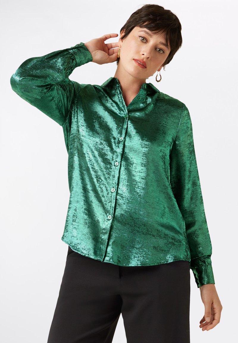 Groene satijnen blouse met een knoopsluiting aan de voorkant, lange mouwen en een klassieke kraag. De stof heeft een glanzende, reflecterende textuur.