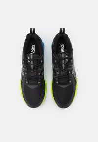 Scarpe sportive nere con tomaia in rete, accenti riflettenti, suola verde e dettaglio del tallone blu. Presentano un sistema di allacciatura e una linguetta posteriore.