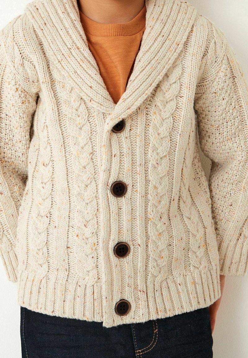 Enfant portant un cardigan crème en maille torsadée avec de gros boutons sur une chemise couleur rouille et un jean foncé.