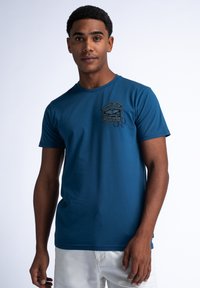 Blauwe katoenen T-shirt met een ronde halslijn, korte mouwen en een reliëflogo op de borst. Model draagt een witte short.