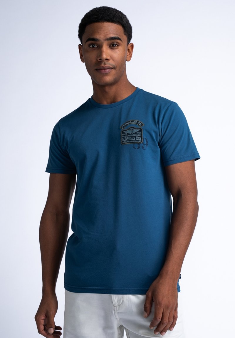 Blauwe katoenen T-shirt met een ronde halslijn, korte mouwen en een reliëflogo op de borst. Model draagt een witte short.