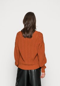 Sweter w kolorze rdzy z dzianiny, z teksturowanym wzorem w zygzaki, luźny krój i ściągacze, zestawiony z czarnymi skórzanymi spodniami.