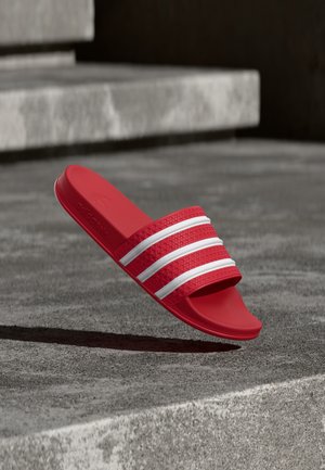 Czerwona sandałka typu slide z trzema białymi paskami spoczywająca na betonowych schodach, z widoczną teksturowaną wkładką i detalami pasków.