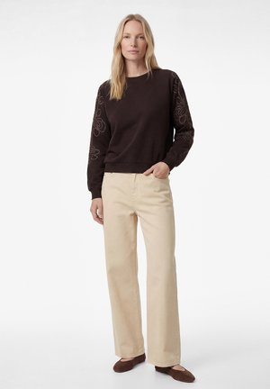 Kvinde med langt blondt hår iført en mørkebrun sweater med blomsterbroderi på ærmerne, beige bukser med vide ben og brune sko.