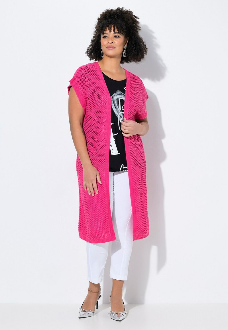 Rosa offen gestrickte Strickjacke über einem schwarzen Grafikshirt, kombiniert mit weißen Hosen und silbernen Absätzen. Verfügt über kurze Ärmel und eine lässige Passform.