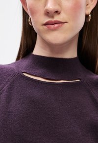 Purpurna pletena majica s visokim izrezom i izrezom, teksturirana tkanina i raglan rukavi, uz niske zlatne hoop naušnice.