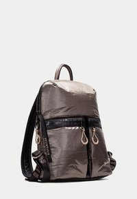 Mochila de tela gris metálico con textura suave. Presenta dos bolsillos frontales con cremallera, correas negras y acentos de logo.