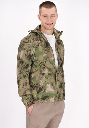 Giacca camouflage in tonalità di verde e marrone, con zip frontale, cappuccio regolabile e tessuto testurizzato. Indossata sopra una camicia bianca.