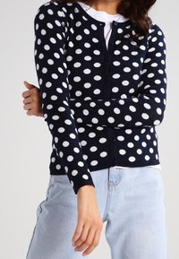 Navy cardigan with white polka dots, round neckline, long sleeves, and button closure, paired with light blue jeans.