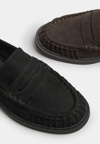 Mocassini in suede neri e marroni presentano dettagli cuciti, tomaia tessurizzata e suole in gomma. Il design include una forma classica di mocassino.