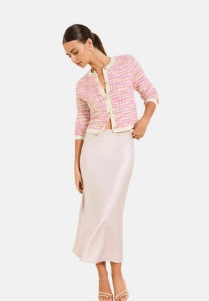 Frau trägt eine rosa-cream karierten Strickjacke mit Knöpfen und einen seidigen, hellrosa Midirock, posiert vor weißem Hintergrund.