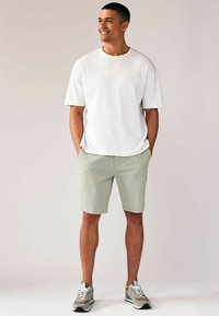 Homme souriant, debout avec les mains dans les poches, portant une chemise blanche à manches courtes, un short vert clair et des baskets grises sur un fond uni.
