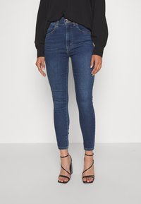 Skinny jeans in dark blue denim, featuring a high waist, five-pocket design, and subtle stitching details, paired with black strappy heels.