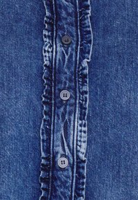 Denimskjorte med en central knapstolpe med fem mørke knapper, flæsedetaljer langs knapstolpen og en middelblå vask.