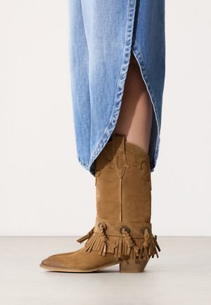 Lichte suède cowboylaars tot halverwege de kuit met franjes en gevlochten details, gedragen met een lichtblauwe denimrok met zijsplit.