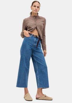 Frau trägt eine kurze braune, geknöpfte Strickjacke, hochtaillierte, weit geschnittene blaue Jeans mit braunem Gürtel und beige Slip-On-Schuhe, steht mit der Hand in der Hosentasche.
