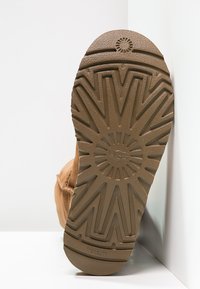 Suola di stivale in gomma color tan con motivo a zigzag testurizzato e emblema circolare, con il logo UGG al centro. Bordo superiore liscio, dettagli minimi.