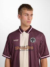 Junger Mann trägt ein dunkelrotes und beige gestreiftes Sportshirt mit den Logos "hummel", "SWARM" und einem runden Emblem, schaut nach links.