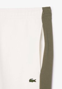 Bavlněné tepláky s bílým tělem a olivově zelenými bočními panely, na levé nohavici je malé vyšívané zelené logo krokodýla.