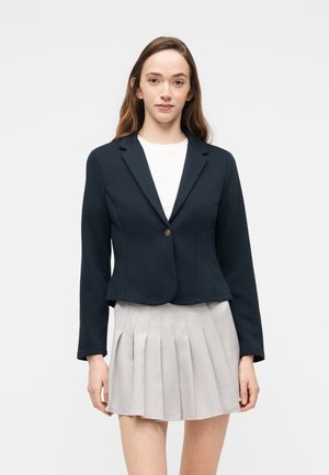 Ung kvinne iført en tettsittende svart blazer over en hvit topp og en lys grå plissert miniskjørt, stående mot en ensfarget bakgrunn.