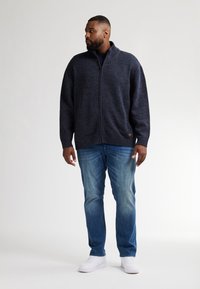 Marinblå zip-up tröja med texturerade axlar, bärs över en mörk skjorta och ljusblå jeans, i kombination med vita sneakers.