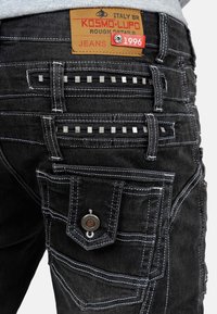 Jeans neri con un'inquadratura ravvicinata delle tasche, cuciture bianche a contrasto, dettagli metallici e una patch in pelle marchiata "KOSMO-LUPO."