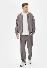 Sudadera gris con cremallera de diseño amplio, combinada con pantalones de chándal grises a juego que tienen rayas laterales, y lleva zapatillas deportivas blancas.
