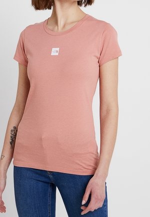 Frau trägt ein eng anliegendes, staubrosa kurzärmeliges T-Shirt mit einem kleinen weißen Logopatch auf der Brust, kombiniert mit blauen Jeans.