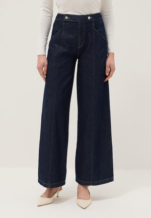 Vrouw draagt donkerblauwe jeans met wijde pijpen en voorste zoomdetails, gecombineerd met een witte top met lange mouwen en beige puntige hakken.