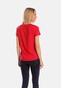 Czerwona koszulka z krótkim rękawem uszyta z miękkiej tkaniny, z okrągłym dekoltem i luźnym fasonem, noszona z dopasowanymi czarnymi dżinsami.