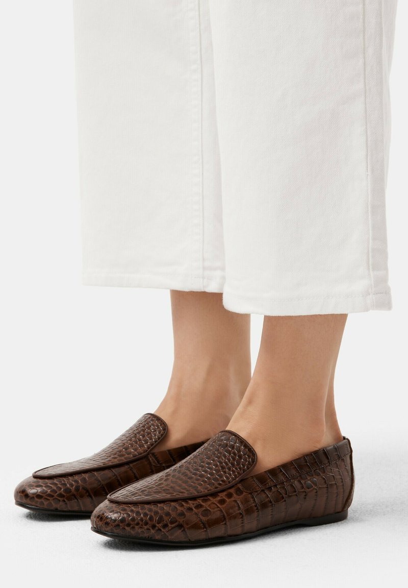 Bruna läderloafers med ett texturerat krokodilmönster, platt sula, rund tå och slip-on design, i kombination med vita byxor.