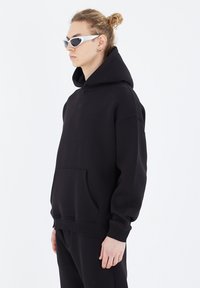 Black hooded sweatshirt made of soft fabric, featuring a front pocket and ribbed cuffs. Worn with narrow, angular sunglasses.