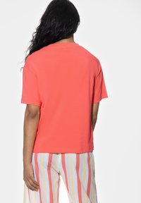 Koralowa koszulka z krótkim rękawem, z okrągłym dekoltem, wykonana z miękkiego materiału, pokazana z tyłu, bez widocznych wzorów ani akcentów.