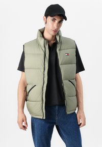 Light olive green puffer vest with a high collar, zipper front, and two side pockets, accented by black trim and a small logo patch.